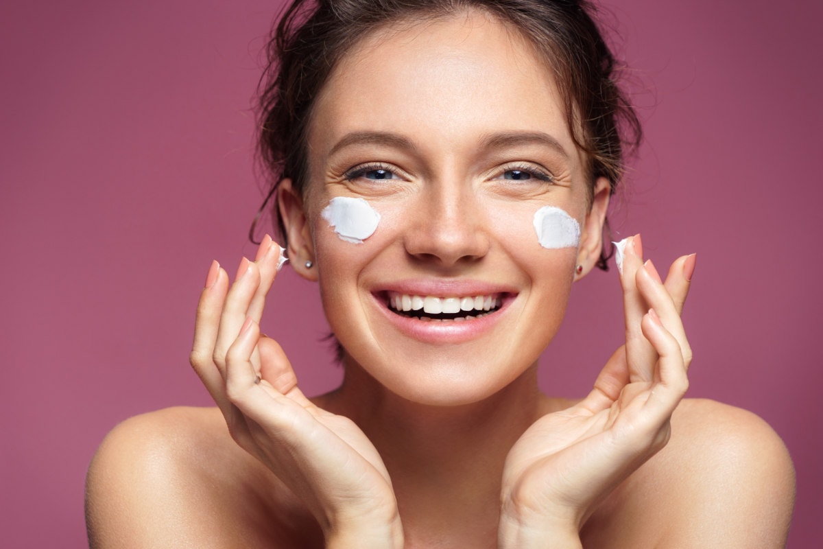 Laughing girl applying moisturizing cream on her face.